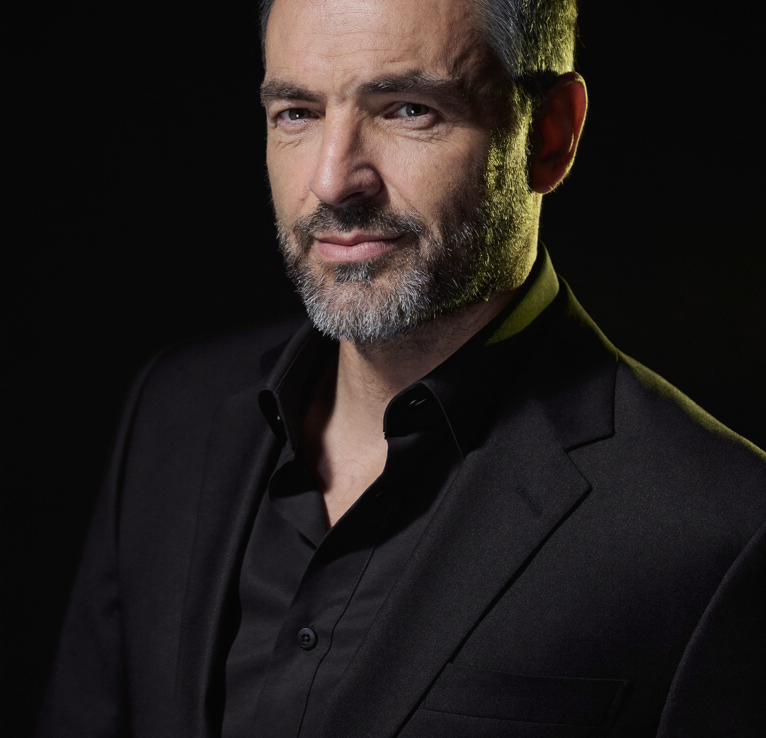 A distinguished, confident professional man in his early fifties in a tailored dark suit, looking rested and composed, lit against a dark background