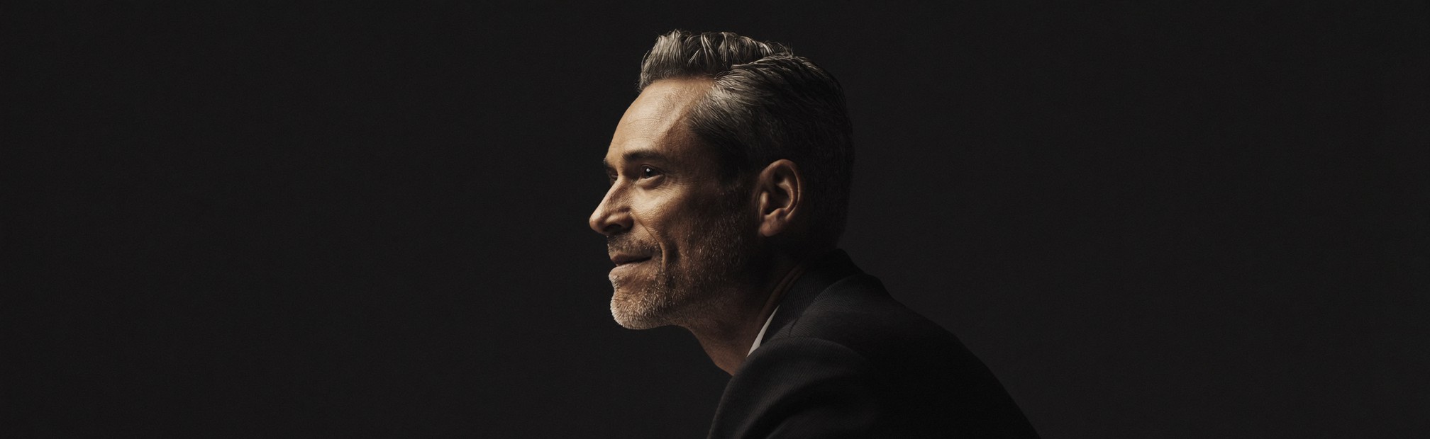A composed professional man in his late forties in three-quarter profile, chin lifted, in dramatic editorial light against a dark background