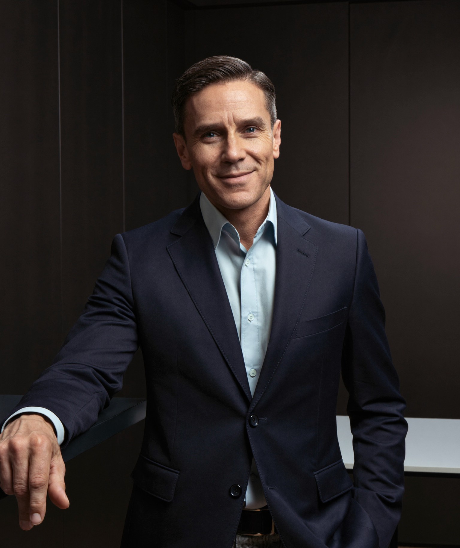 A board-certified facial-plastic surgeon in a tailored navy blazer, standing in a modern consultation office